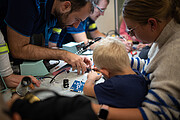 A young child is seen from the back, working on a printed circuit board, being helped by an ESO engineer.