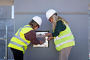 Two people in high-visibility jackets and hard hats are standing on either side of a shiny, metallic square box – the time capsule – placing it into a grey concrete wall.