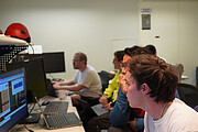 Das Foto zeigt eine Gruppe von Personen in einem Kontrollraum des Paranal-Observatoriums. Sie sitzen konzentriert an mehreren Computern und betrachten Bildschirme mit wissenschaftlichen Daten und Diagrammen. Im Vordergrund ist eine Person im Profil gut zu erkennen, die intensiv auf den Monitor blickt. Dahinter sitzen weitere Personen nebeneinander in einer Reihe, ebenfalls auf die Bildschirme fokussiert. Die Atmosphäre wirkt ruhig und konzentriert. Im Hintergrund sind schlichte, helle Wände, einige technische Geräte und rote Schutzhelme zu sehen. Das Bild vermittelt den Moment der ersten Auswertung von Beobachtungsdaten des 4MOST-Instruments.