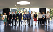 Eine Gruppe von Personen steht in einem modernen, lichtdurchfluteten Raum mit großen Fenstern und Blick ins Grüne. Die Personen sind in einer Reihe aufgestellt und blicken zur Kamera. In der Mitte tragen mehrere Anwesende Anzüge, während andere formell oder business-casual gekleidet sind. Über der Gruppe hängt ein großer, runder Deckenleuchter. Im Hintergrund ist eine Fahne mit dem ESO-Logo zu erkennen. Die Szene vermittelt den Eindruck eines offiziellen Anlasses, passend zum Kontext einer Vertragsunterzeichnung der ESO im Jahr 2026.