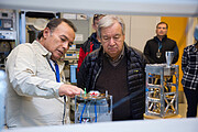 In the foreground of the image, two men look closely at a small metallic structure with a hexagonal top plate. The man on the left is resting his hand on the device. To the right is another metallic structure, and the image is taken within a laboratory, where three other people are standing within frame in the background.