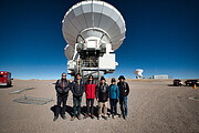 The image shows a desert landscape under blue skies. Six people wearing differently coloured jackets are facing the camera. Right behind them stands a large white antenna, with other similar ones in the distance.