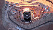 In the middle of the image is the dome of the telescope from a top-down perspective. The dome has a rectangular hole from the bottom to two-thirds the way up, on either side are the beams that make up the skeleton structure of the doors. Around the dome is red desert with cranes, shipping containers and a winding road.