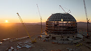 This image shows a large, metallic dome on top of a mountain, against the landscape of the Chilean desert at sunset. There are cranes and other construction equipment around. Near the base of the dome there's the tiny figure of a man in a reflective jacket.