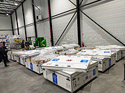 In a warehouse setting, several white, large containers with the ESO logo — four stars around the letters E, S, O on a blue background — sit on a concrete floor. Some of the boxes have open lids with plastic sheets sticking out. Behind them, to the back left of the image, a few people stand around. A large white wall with black supports fills the top half of the image.