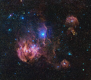 Wispy pink clouds pop out of a dark background. The brightest and largest cloud is offset from centre to the lower left. A cloud ridge cuts vertically through the centre of the frame, with a large bright blue-white spot in the middle. To the right of the image are three smaller clouds. Dotted across the image are blue, orange and white points of varying size and brightness.