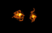 This is a telescopic image of two galaxies, shown as bright clouds of swirling gas. Against the pitch-dark backdrop of deep space, these two blotchy clouds take up most of the picture, and they are shown in bright yellows and oranges that get more intense at their centres and fade towards their edges.