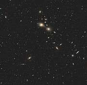 This image shows a large patch of space, dotted with small, distant stars and broken up by fuzzy galaxies spread throughout. These warm, yellow-ish smears of light sit at all angles, some looking more like spirals and some more like clouds. A few galaxies are closer together than others. At the centre of the image is a tiny white dot, which is where the two jousting galaxies can be found.