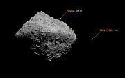 This image shows a size comparison between two asteroids. The background is completely black, with the larger of the two asteroids taking up the left side of the image and spilling into the right half. It is shaped like a rough diamond, with small rocks sticking out of the surface. There is a label with the words “Ryugu - 900m