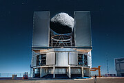 The image shows the size comparison between a telescope inside a cylindrical dome and an asteroid. The telescope is at the centre of the image, taking up more than a third of the image horizontally. The asteroid fits between the open doors of the telescope’s dome, where it sits above the main structure that holds the telescope's mirrors.