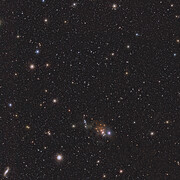 A wide telescopic view of a starry field. There are countless faint stars and a few bright ones in shades of orange, blue and white. At the bottom centre there is a small orange wispy cloud.