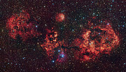 A telescopic view of a cosmic cloud against a dark, starry sky. The cloud resembles a flying bat, spanning its wings. The cloud is mostly red, with some areas partially blocked by dark dust clumps.
