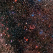 A wide view of a region of the night sky full of stars covered by a reddish glow. At the centre there is a small group of clouds that resemble a flying bat spanning its wings.