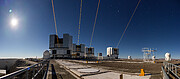 This photograph was taken at night, but with an exposure time giving the impression it’s daytime. The Moon is shining brightly in the top left corner, with the blue illuminated night sky in the background. In the centre of the image are the four Unit Telescopes, which are large, cylindrical, grey domes. An orange beam emerges from each of these telescopes, piercing the sky at a point that appears to be behind the person who took the photograph. The site where the Unit Telescopes are located appears bulky, with some other smaller white telescopes distributed around it.