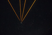 This image shows the black night sky, with some differently bright stars, resembling white paint splatters. Four orange beams, coming from the top of the image, converge at a single spot that is slightly to the left of the centre. A small grey speckle is present at this spot, which is the observation target.