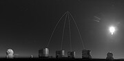 This is a panoramic 360-degree spherical view of the night sky from an observatory, captured in black and white. In the top right part, six cylindrical domes are aligned along the panoramic circle, which represents the ground. The Moon shines bright in the top-right inner part of the circle. The four largest domes are the Unit Telescopes, each of which has a beam emerging from it. These four beams then meet at one point in the sky in the lower centre of the image.
