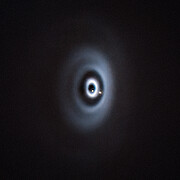A telescopic image of a flat, inclined fuzzy disc with bright rings and dark gaps, all concentric. There is a small blob in one of those gaps. An inset that zooms in on the centre of the disc shows a second blob in another gap.