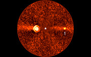 Runde, rötlich gefärbte Aufnahme eines Himmelsausschnitts, die mit astronomischen Instrumenten erstellt wurde. Im Zentrum befindet sich ein heller, kreisförmiger Bereich, der von einem Sternsymbol markiert ist. Rechts davon ist ein kleiner, leuchtender Punkt zu sehen, auf den ein weißer Pfeil zeigt. Die gesamte Fläche ist von körnigen Strukturen und einem horizontal verlaufenden, helleren Streifen durchzogen. Die Bildgestaltung und Markierungen deuten auf die Darstellung eines Exoplaneten in unmittelbarer Nähe zu seinem Zentralstern hin.