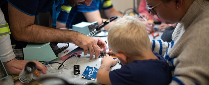 A young child is seen from the back, working on a printed circuit board, being helped by an ESO engineer.