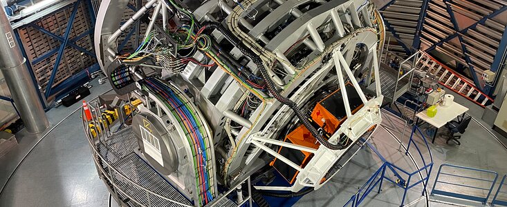 Das Foto zeigt das VISTA-Teleskop der Europäischen Südsternwarte (ESO) in seiner Kuppel am Paranal-Observatorium in Chile. Das große, silbern-graue Teleskop dominiert den Raum; es steht auf einer drehbaren Plattform und ist von Geländern, Kabeln und technischen Geräten umgeben. Deutlich sichtbar ist der neu installierte 4MOST-Spektrograf – ein komplexes Instrument mit zahlreichen bunten Kabeln, Rohren und weißen Stützstrukturen, die sich um die Rückseite des Teleskops winden. Zwei orangefarbene Elemente markieren den Bereich, in dem 4MOST befestigt ist. Im Hintergrund sind die metallisch-blauen Streben der Kuppel sowie Arbeitstische mit Werkzeugen und Computern zu sehen, die auf laufende technische Tests hindeuten. Die Szene vermittelt den Eindruck einer hochmodernen astronomischen Anlage während der erfolgreichen Inbetriebnahme eines neuen wissenschaftlichen Instruments.