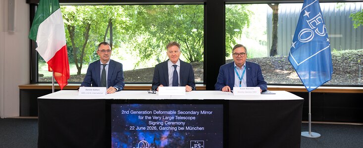 Drei Männer sitzen an einem Tisch, der mit einem weißen Tuch bedeckt ist. Vor ihnen steht ein großes Schild mit englischsprachigem Text, das auf die Unterzeichnung eines Vertrags für den „2nd Generation Deformable Secondary Mirror for the Very Large Telescope“ am 22. Juni 2026 in Garching bei München hinweist. Links steht eine italienische Flagge, rechts eine blaue Flagge mit dem ESO-Logo. Im Hintergrund sind große Fenster mit Blick auf grüne Bäume und eine Rasenfläche zu sehen. Die Szene vermittelt den offiziellen Charakter einer Vertragsunterzeichnung zwischen Vertretern der ESO und weiteren Beteiligten.