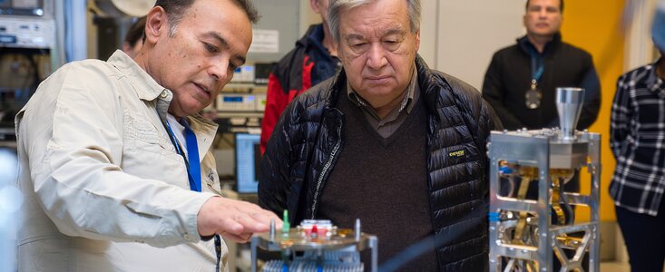 In the foreground of the image, two men look closely at a small metallic structure with a hexagonal top plate. The man on the left is resting his hand on the device. To the right is another metallic structure, and the image is taken within a laboratory, where three other people are standing within frame in the background.