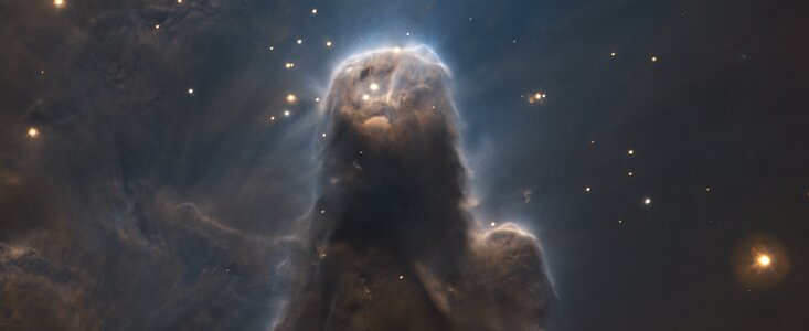 The image shows the Cone Nebula at the centre of the picture taking up most of the frame. The shape of the dark nebulous cloud is that of a cone, pointing upwards. At the top and surrounding the cone are bright yellow stars of different sizes, tiny dots and bigger gassy spheres. To the left of the central figure, abstract dusty patterns are fading upwards. The top shines brightly, lit up by luminous stars.