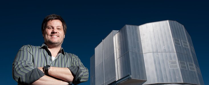 Ein Mann steht mit verschränkten Armen vor einem großen, silbrig glänzenden Teleskopgebäude unter tiefblauem Himmel. Er lächelt und blickt in die Kamera. Das Teleskop hat eine kantige Metallfassade und dominiert den rechten Bildrand, während der Mann links im Vordergrund steht. Das Bild vermittelt einen klaren, sonnigen Tag an einem Observatorium.