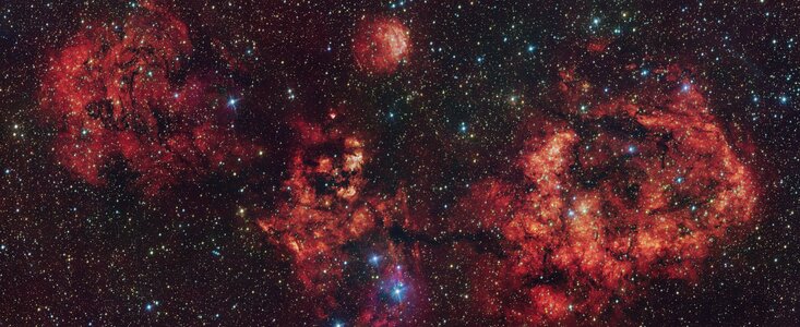 A telescopic view of a cosmic cloud against a dark starry sky. The cloud resembles a flying bat, spanning its wings. The cloud is mostly red, with some areas partially blocked by dark dust clumps.
