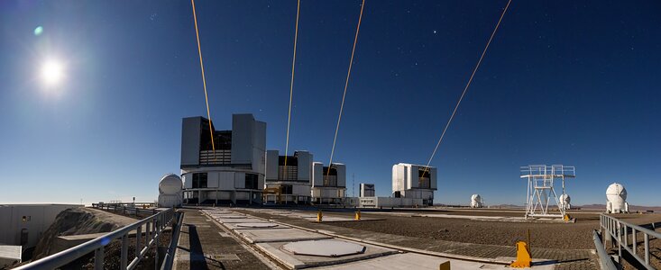 This photograph was taken at night, but with an exposure time giving the impression it’s daytime. The Moon is shining brightly in the top left corner, with the blue illuminated night sky in the background. In the centre of the image are the four Unit Telescopes, which are large, cylindrical, grey domes. An orange beam emerges from each of these telescopes, piercing the sky at a point that appears to be behind the person who took the photograph. The site where the Unit Telescopes are located appears bulky, with some other smaller white telescopes distributed around it.