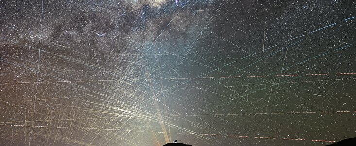 Eine weitwinklige Nachtaufnahme der Atacama-Wüste unter einem von unzähligen Satellitenspuren durchzogenen Sternenhimmel. Am unteren Bildrand erstreckt sich die dunkle Silhouette einer bergigen Wüstenlandschaft. Zentral auf der Spitze des höchsten Hügels im Vordergrund steht die beleuchtete, kuppelförmige Baustelle des Extremely Large Telescope (ELT).

Vom Horizont weit hinter dem Hügel ragen vier dünne, orangefarbene Laserstrahlen des Paranal-Observatoriums vertikal in den Himmel. Der obere Teil des Bildes wird vom bandförmigen, detailreichen Leuchten der Milchstraße dominiert, die sich diagonal von links nach rechts erstreckt und von dunklen Staubwolken durchsetzt ist.

Das gesamte Himmelszelt ist von einem dichten, chaotischen Netz aus Hunderten geradlinigen Lichtspuren überzogen. Diese Linien verlaufen in alle Richtungen und kreuzen sich vielfach. Besonders im linken unteren Quadranten, nahe dem Horizont, ist die Dichte der weißlichen Satellitenspuren so extrem, dass sie ein fast geschlossenes, helles Geflecht bilden, das den Blick auf die Sterne dahinter blockiert. Einige wenige, horizontal verlaufende Spuren im rechten Bildbereich bestehen aus rot-weiß gestrichelten Linien, die von den Blinklichtern vorbeifliegender Flugzeuge stammen.