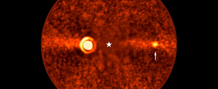 Runde, rötlich gefärbte Aufnahme eines Himmelsausschnitts, die mit astronomischen Instrumenten erstellt wurde. Im Zentrum befindet sich ein heller, kreisförmiger Bereich, der von einem Sternsymbol markiert ist. Rechts davon ist ein kleiner, leuchtender Punkt zu sehen, auf den ein weißer Pfeil zeigt. Die gesamte Fläche ist von körnigen Strukturen und einem horizontal verlaufenden, helleren Streifen durchzogen. Die Bildgestaltung und Markierungen deuten auf die Darstellung eines Exoplaneten in unmittelbarer Nähe zu seinem Zentralstern hin.