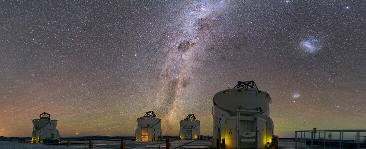 A landscape picture of the VLT's four auxiliary telescopes against the vast background of the night sky. The Milky Way, in the centre of this image, is pointing upwards and is surrounded by countless stars. The Large Magellanic Clouds are also visible. Towards the horizon in the sky, a glow of orange and green appears.