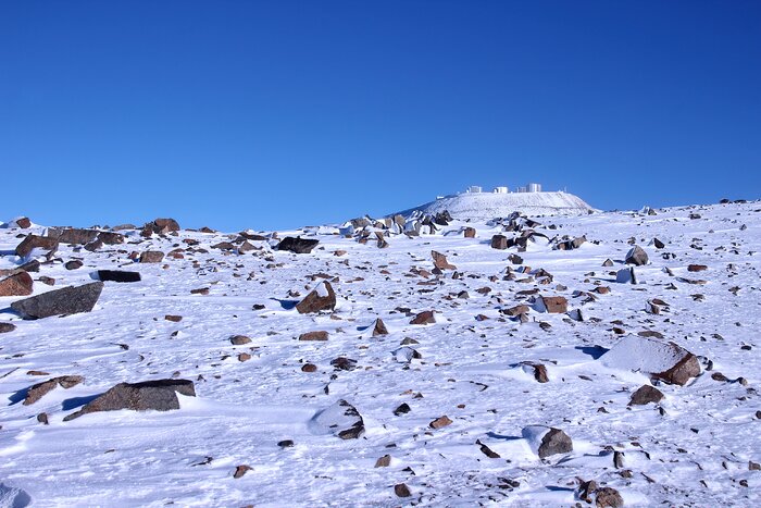 A rocky mountain slope covered by a thin layer of snow, with dark brown rocks scattered across the foreground. At the top of the slope, several white and silver observatory buildings with cylindrical telescope domes stand against a deep blue, cloudless sky.