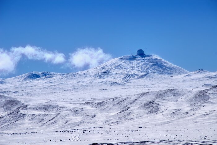 A broad mountain landscape covered in white snow, with darker rocky ridges and shallow tracks visible across the slopes. A large, rounded telescope dome stands alone on the summit near the centre of the image, while a low white cloud drifts behind the mountain under a deep blue sky.