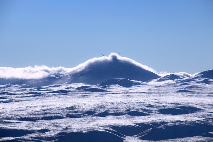 A snow-covered landscape of rounded hills stretches towards a dark mountains in the distance. Low clouds curl over the summit, partly obscuring a large round dome silhouetted against a pale blue sky.