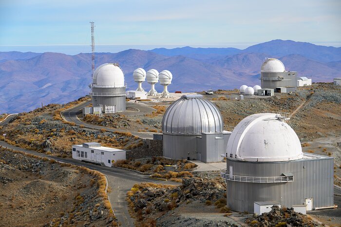 This landscape image shows a landscape with four cylindrical-shaped buildings with hemispherical domes, three identical structures made of white sphere on square-based pyramid-shaped supports, and several smaller white cuboid-shaped buildings. All the domes are white except the one second from the front which has a shiny silver dome. Roads snake between the buildings, and the land between is dotted with orange-brown bushes. In the distance are reddish-blue mountains against a pale blue sky.