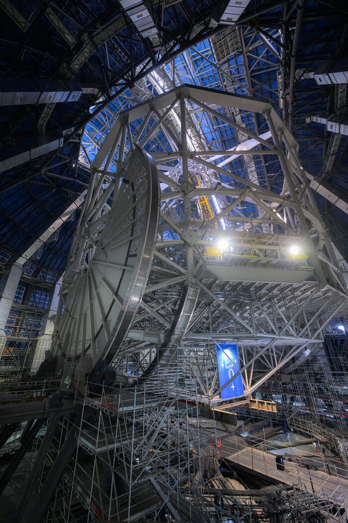 This image in portrait shows a metal structure above, composed of many beams, with scaffolding and a blue sign with the ESO logo below. A half-circle shape in metal is on the left-hand side of the metal structure.