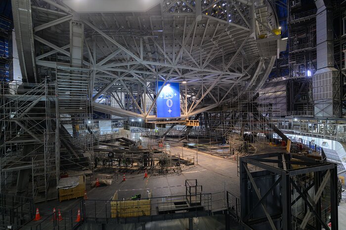 This landscape image shows a large space with a metal structure made of beams with a blue sign with the ESO logo and scaffolding below it. Orange cones each with two white stripes are scattered around the floor and reveal the gigantic scale of the central structure.