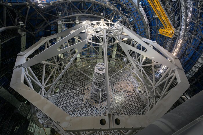 This landscape image shows a metal structure from above. There is circular shape at the base with a tall metal post, joined to surrounding metal beams by five long horizontal metal supports. There is a twelve-sided metal structure at the top, with a gap in its centre, which the surrounding beams attach to.