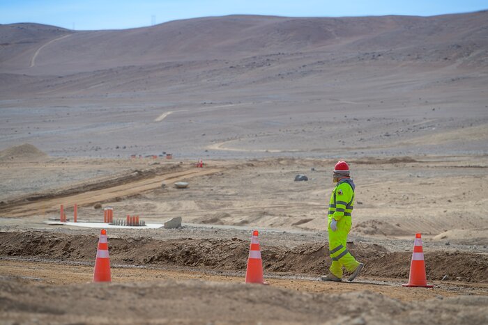 This landscape image shows brown ground carved with lines and mounds of dirt, with reddish-brown hills in the background, above which is a slither of pale blue sky. In the foreground, on the right, a person in a fluorescent yellow vest, red hard hat, gloves, and a face covering walks facing left. In front of them are three equally spaced orange cones, each with two white stripes.