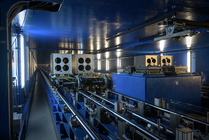 This landscape image shows a wide corridor with blue ceiling and walls. There is a walkway next to the left-hand wall, vertical white lights along both walls, and shiny metal tracks on top of blue metal in the centre and on the right. On these tracks are four cube-shaped objects with wheels, each with four black circles on the objects’ front face.