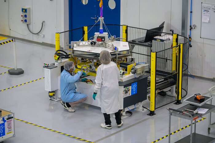 This landscape image shows a technical facility with light grey floors marked with yellow and black tape, a light grey wall to the back with blue double doors on the left hand side, and a structure at the centre with a hexagonal-shaped object held above it by a central rope and support and a black metal mesh with yellow vertical supports around it. Two people wearing hairnets are facing the structure: one on the left wearing a light blue labcoat, crouching and turning a shiny metal wheel on the structure, and one on the right standing in a white labcoat.