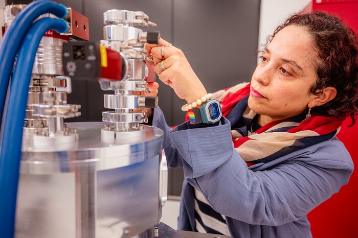 The image is a close-up picture of a woman with short, curly and dark red hair, examine a technological instrument. The instrument has a shiny, metallic colour and connects to a pair of striking red blue tubes.
