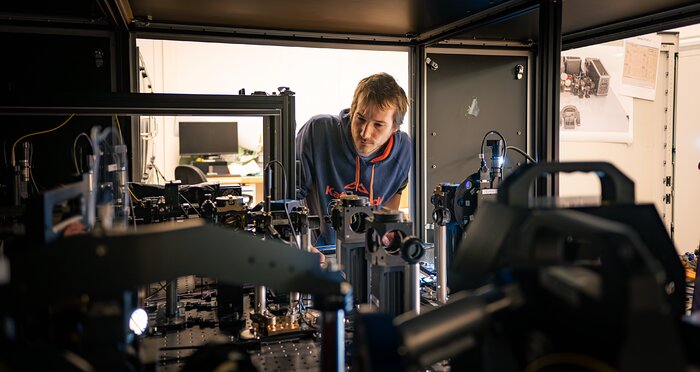 A man with shourt brown hair wearing a blue hoodie is seen across some technological instruments, inspecting something in his vicinity. These instruments appear to be fitted inside a black structure slightly taller than a regular person's height.