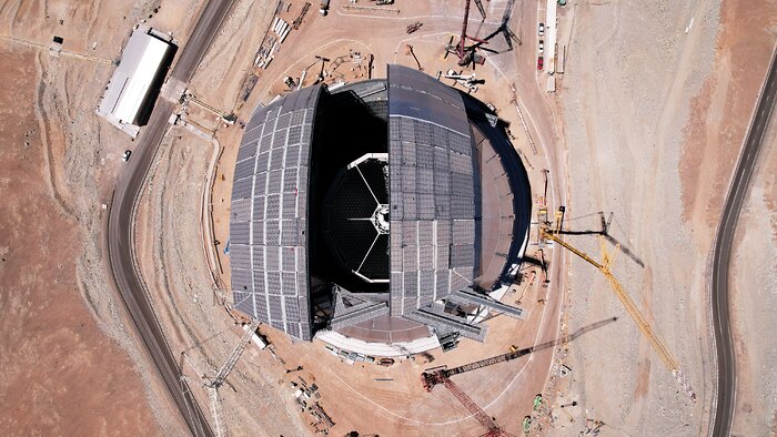 This drone shot is a view from above of the Extremely Large Telescope with one of its slit doors open and other closed, and cranes surround the central telescope. Half of a spider-like frame structure is visible through the open door. Roads circle the land below the ELT and the land is shades of orange-brown.