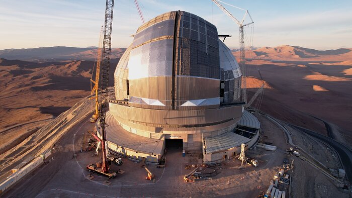 At the centre of the drone image, the ELT construction site stands out against the vast expanses of the Atacama desert, with surrounding orange hills shown with deep shadows. Its giant closed dome is covered in shiny metal corrugated plates. Giant cranes are around the building, underlining that construction is not finished.