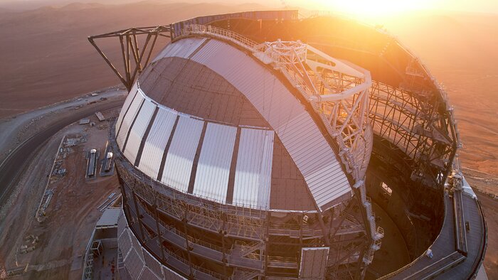 This aerial photo shows the top of the Extremely Large Telescope. Its dome is still under construction and remains open. Some plates of aluminium have already been set and cover a small section of the dome's outer surface. Due to this, the orange light of the sunset goes through the dome, illuminating the main structure of the telescope. This is a cage-like structure, 50-metre tall, mostly made of white-coloured steel. The background shows the barren landscape of the Atacama Desert.