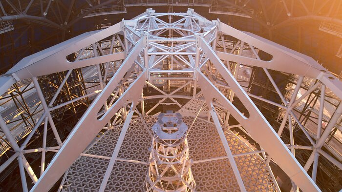 This drone footage shows a close-up of the main steel frame inside the Extremely Large Telescope. The shape of the frame is a decagonal prism, with four main single frames attached at the top into a smaller hexagonal frame. Below stands the 50-m tower. At the base, there is a honeycomb-like structure. Around this main frame appears the interior of the ELT's dome.