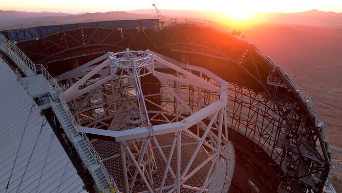 This drone image shows the inner section of the Extremely Large Telescope. The sunset's orange light is glaring over the dome, parts of its metal frame are showing. Inside the dome stands the telescope's altitude structure. This large steel frame looks like a cage, with a shape of a decagonal prism. Attached to this frame is a smaller, yet still large semicircular steel plate, which is perforated.